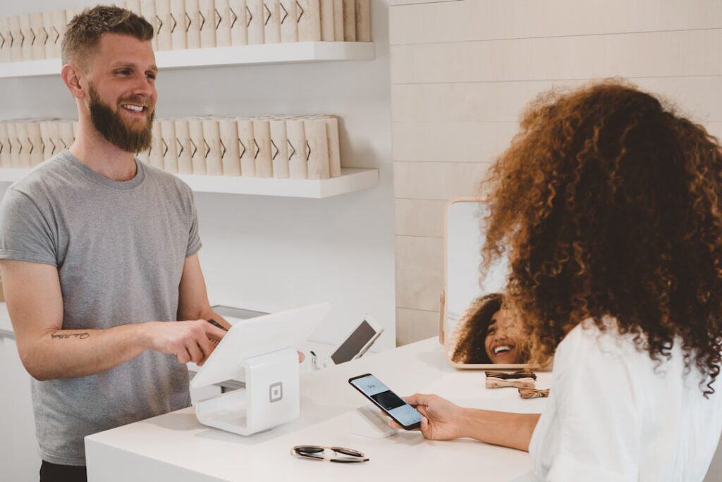 A person with curly hair makes a payment using a smartphone at a modern store counter, while the bearded cashier smiles. Sunglasses rest on the white countertop.